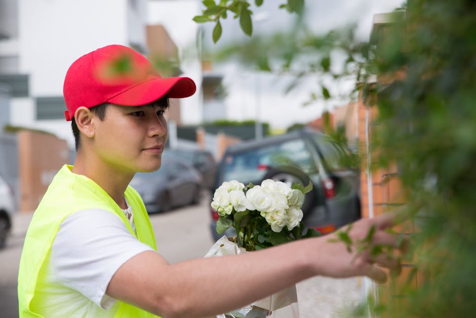 A young delivery person wearing a red cap and high-visibility yellow vest is outdoors, holding a bouquet of fresh white roses wrapped in paper. They are reaching out to attach or adjust the floral gif