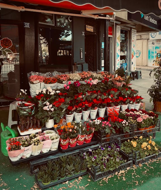 An outdoor display of fresh flowers arranged in white, red, and pink wrapping paper and plastic containers, set on black pallet racks outside a flower shop. The bouquet includes roses, carnations, and