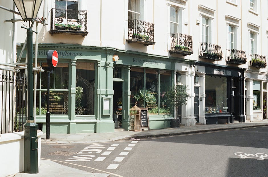 A row of high street shops featuring a flower shop with a pastel green facade and large glass windows displaying fresh floral arrangements. The shop is labeled 'Kensington Flowers' and has a small cha