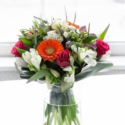 This lush floral composition, thoughtfully designed in the heart of Thornton Heath, is a testament to the harmony of colour, texture, and emotion that defines expert floristry. The bouquet's gentle halo of creamy peach and beige gerbera daisies encircles the striking orange centers, imparting a sense of joy akin to spring's first dawn at nearby Norbury Hall Park. Deep, velvety magenta roses lend a sumptuous depth and romantic richness, while clusters of ivory alstroemeria and whisper-light sprays of small white blossoms bring brightness and a tender vibrancy. Generous layers of glossy, paddle-shaped aspidistra leaves enfold the base, grounding the blooms in calm, leafy serenity and enhancing their rounded, lush silhouette. Slender pointed greenery and sprigs of delicate foliage interweave throughout, adding detail and visual rhythm. Each flower is illuminated by soft, diffused light, enhancing the spectrum of hues without diminishing the arrangement's natural beauty. Displayed in a tall, crystal-clear vase that reveals the vividly green stems, the bouquet conveys freshness and a sense of new beginnings-perfect for a gathering of friends, a congratulatory toast, or a thoughtful gift for someone embarking on a new chapter in Thornton Heath. Every detail speaks of optimism, care, and the local artistry that defines arrangements made for and inspired by the Thornton Heath community.